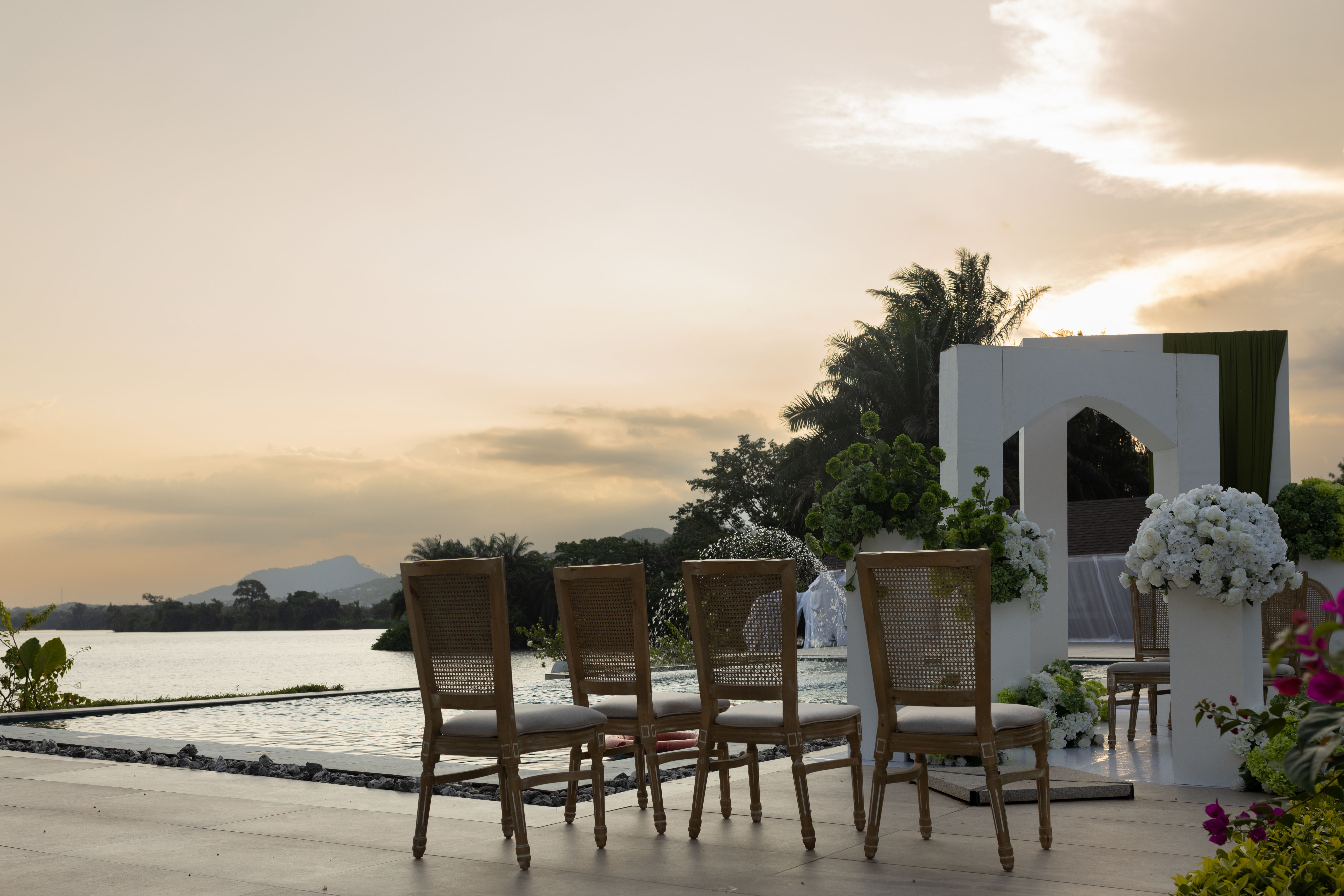 Wedding ceremony setup at Bridgeview Resort in Akosombo, Ghana, overlooking the Volta Lake, with wooden chairs, white floral arrangements, a modern wedding arch, and a sunset backdrop.
